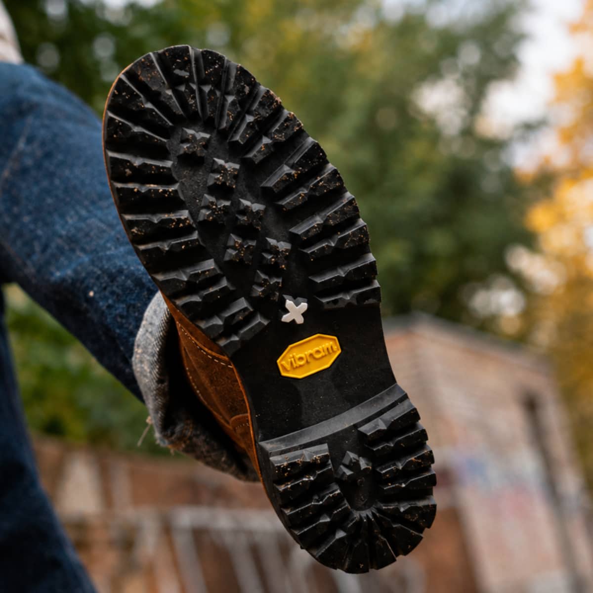 A close-up shot of a Vibram boot sole being held up by a person wearing blue jeans, with an outdoor blurred background.
