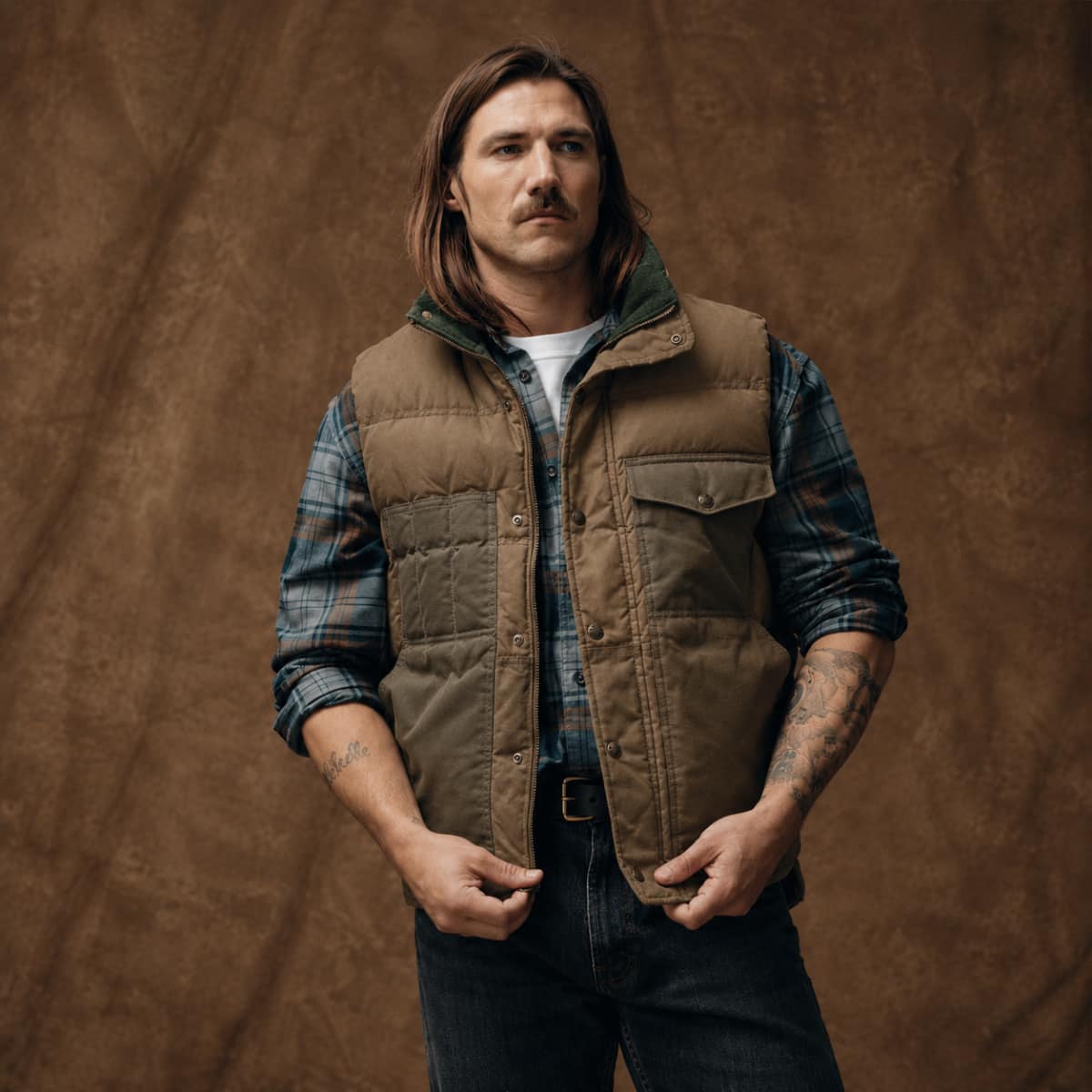 A man wearing a brown Filson Down Cruiser Vest over a blue plaid shirt and dark jeans, standing against a textured brown studio backdrop.