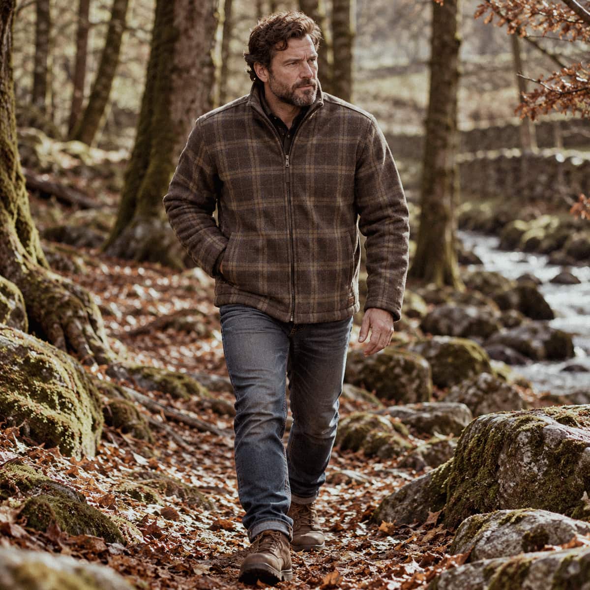 A man walking along a rocky forest path wearing a brown and black plaid zip-up wool jacket, blue jeans, and brown boots.