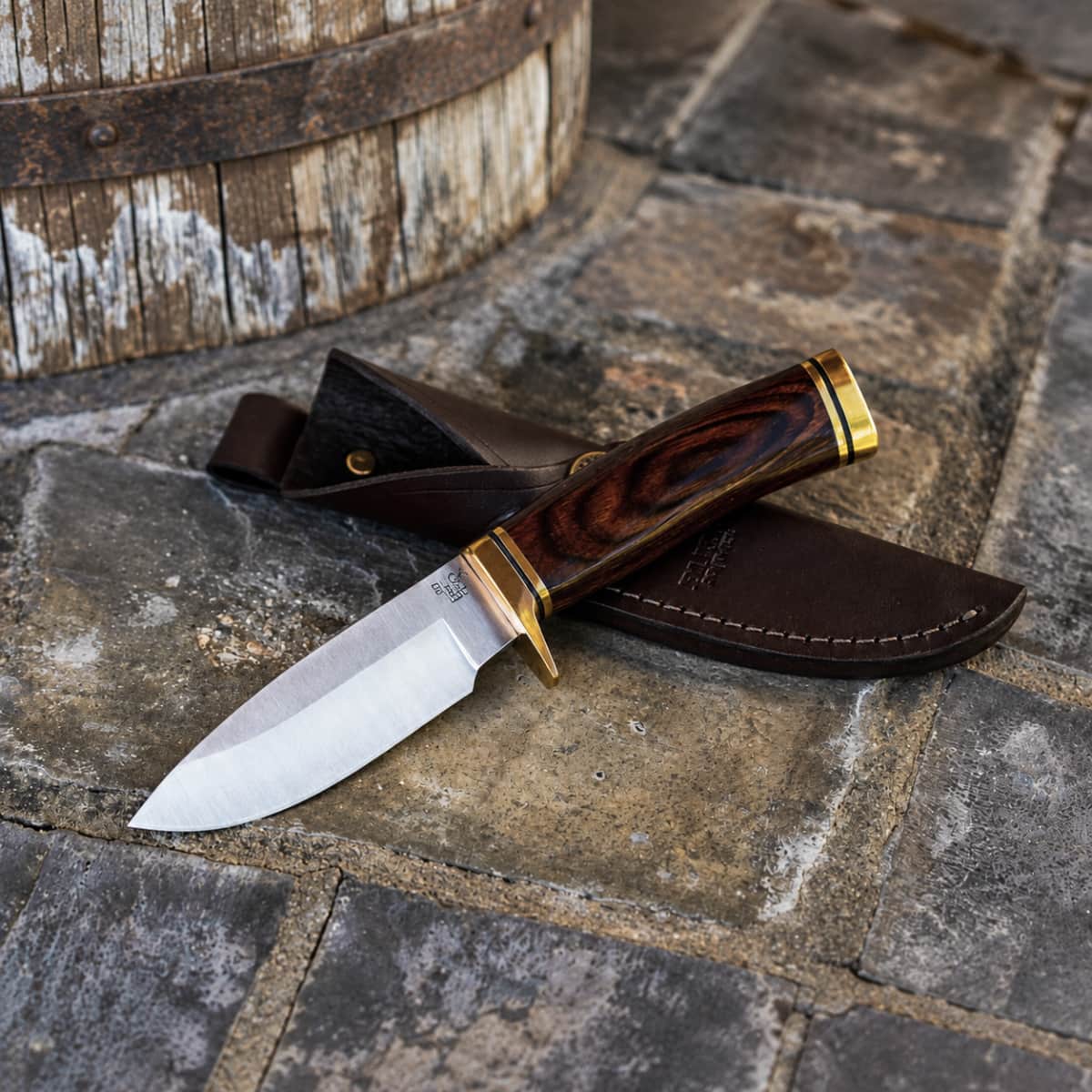 A fixed-blade knife with a polished steel blade, dark wood handle, and brass accents rests on a dark brown leather sheath, placed on a stone surface next to a wooden barrel.