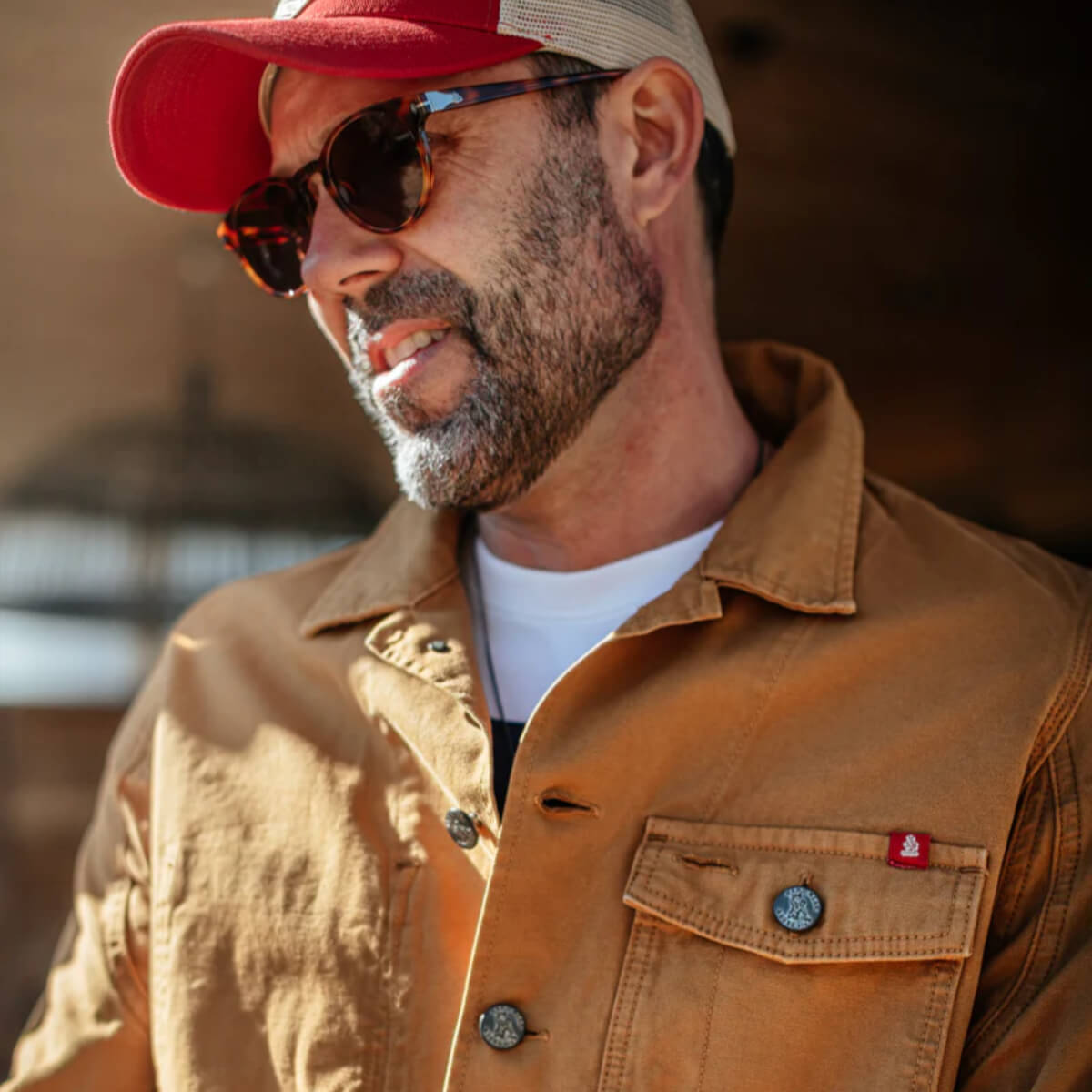 Man wearing a red cap, sunglasses, and a brown jacket with a blurred background