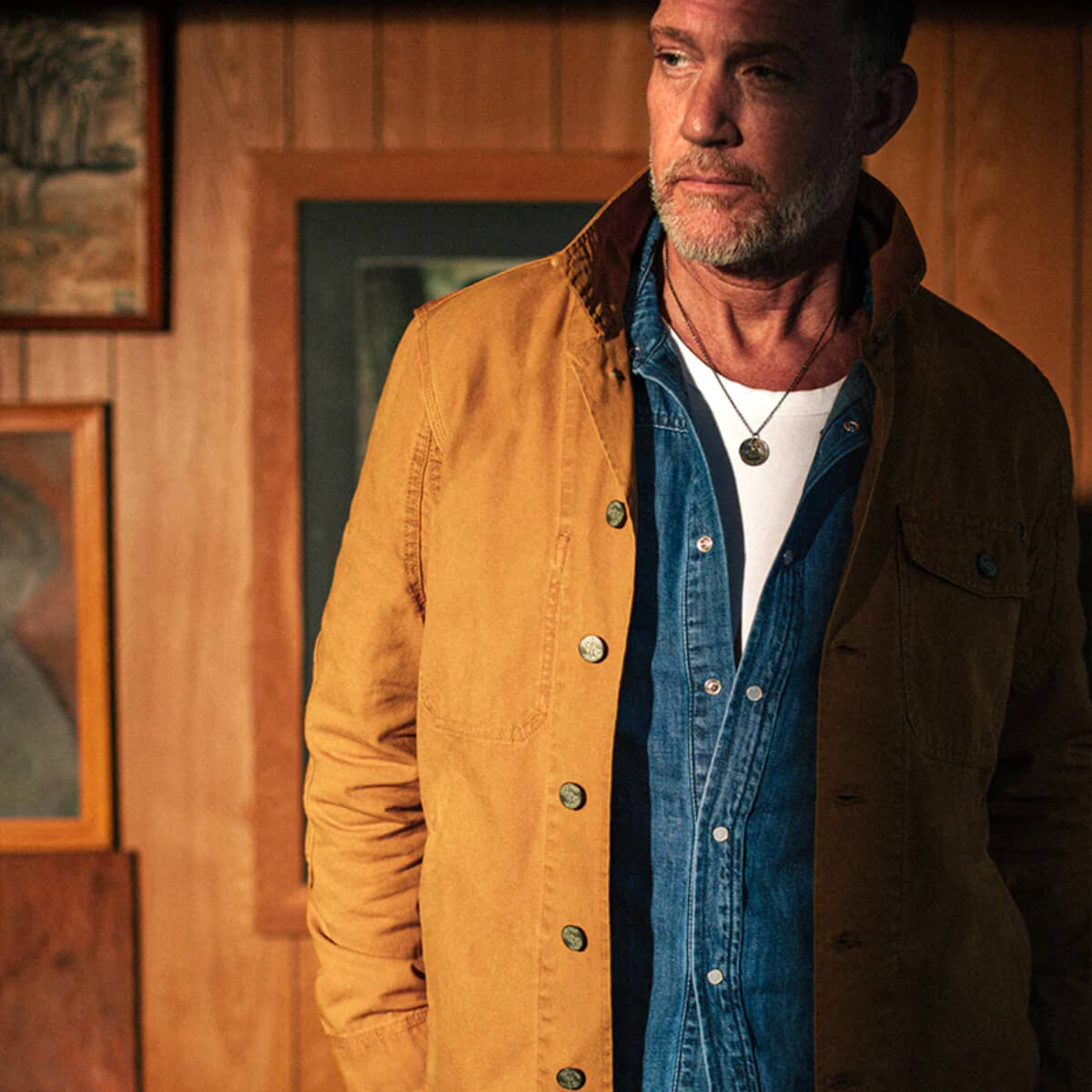 Man wearing a mustard yellow jacket over a denim shirt in a rustic setting