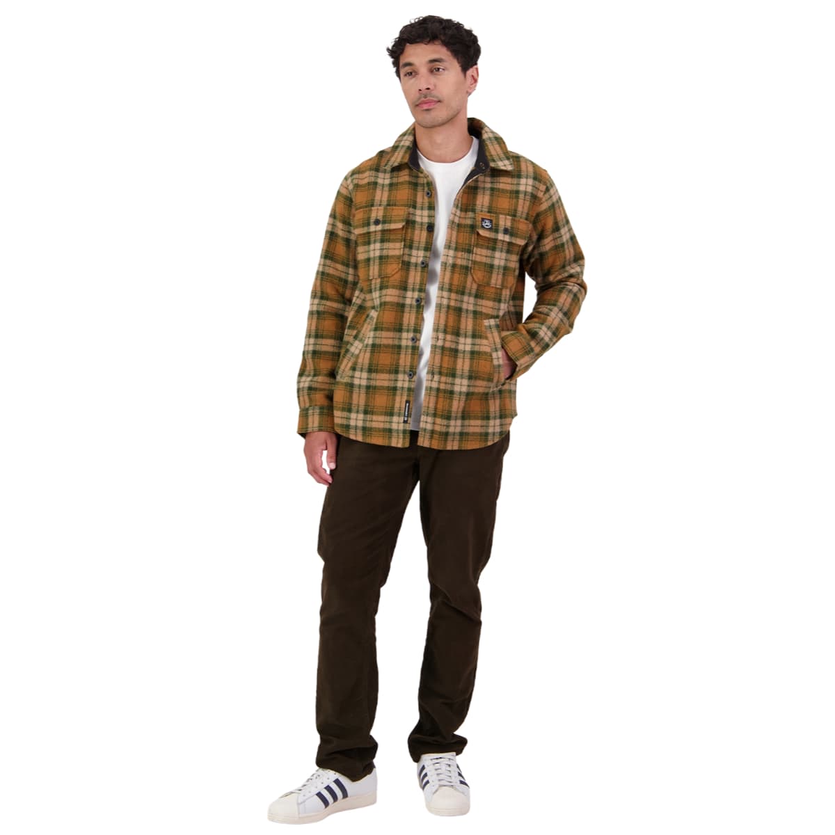 A man modeling a brown and yellow plaid overshirt with a white t-shirt, dark brown pants, and white sneakers, photographed against a solid white background.