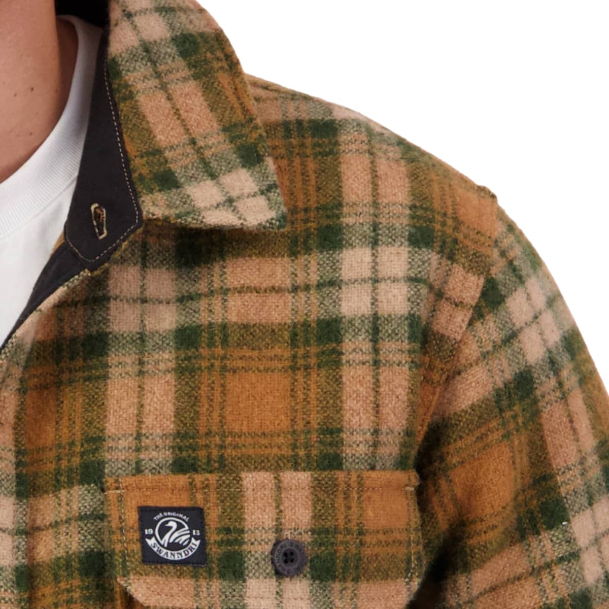 Close-up of a person wearing a plaid overshirt with a tan, green, and cream pattern, featuring a chest pocket with a visible 'Wanderers' brand patch.