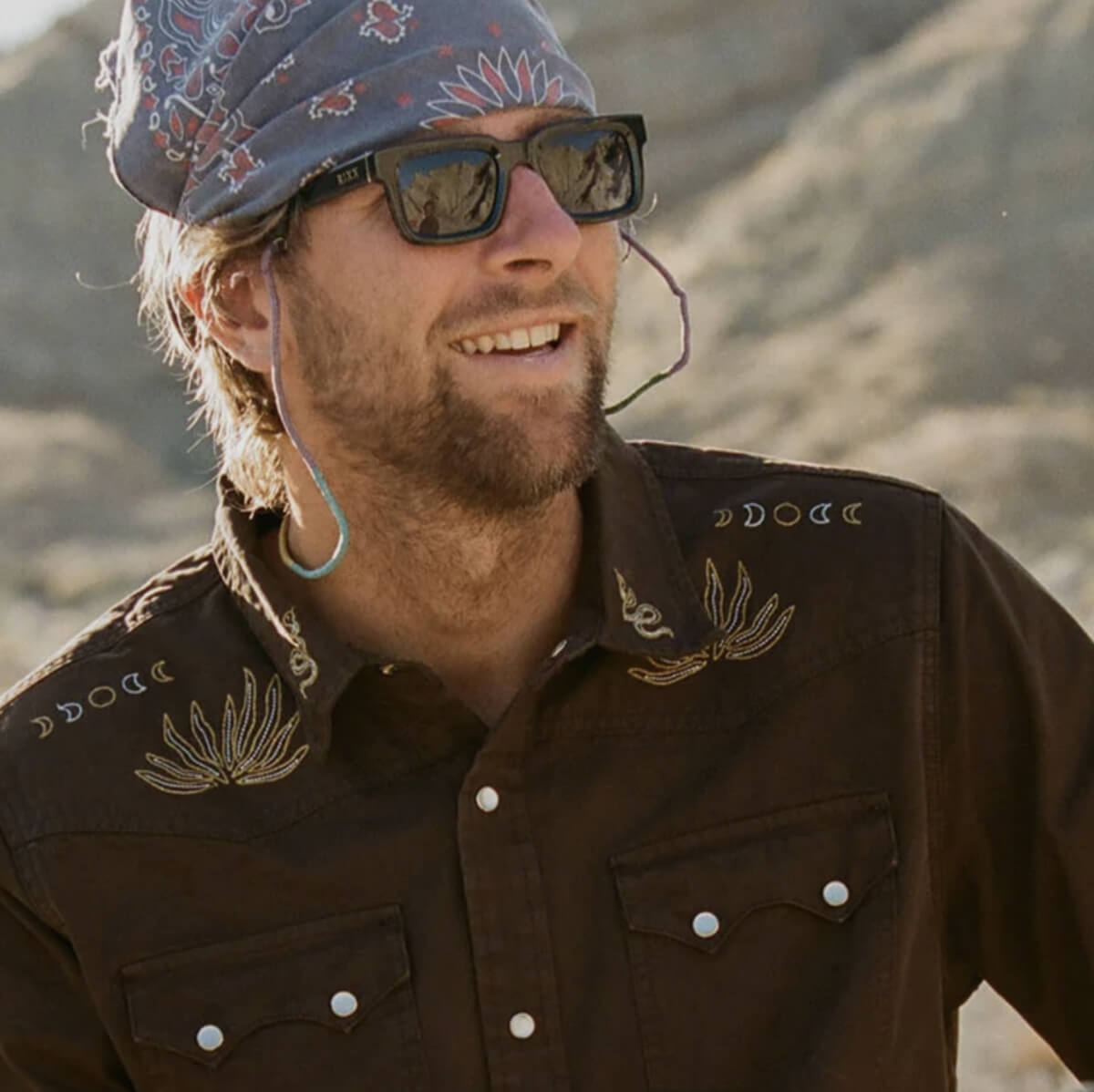 Man wearing a brown embroidered shirt and bandana with desert landscape in the background