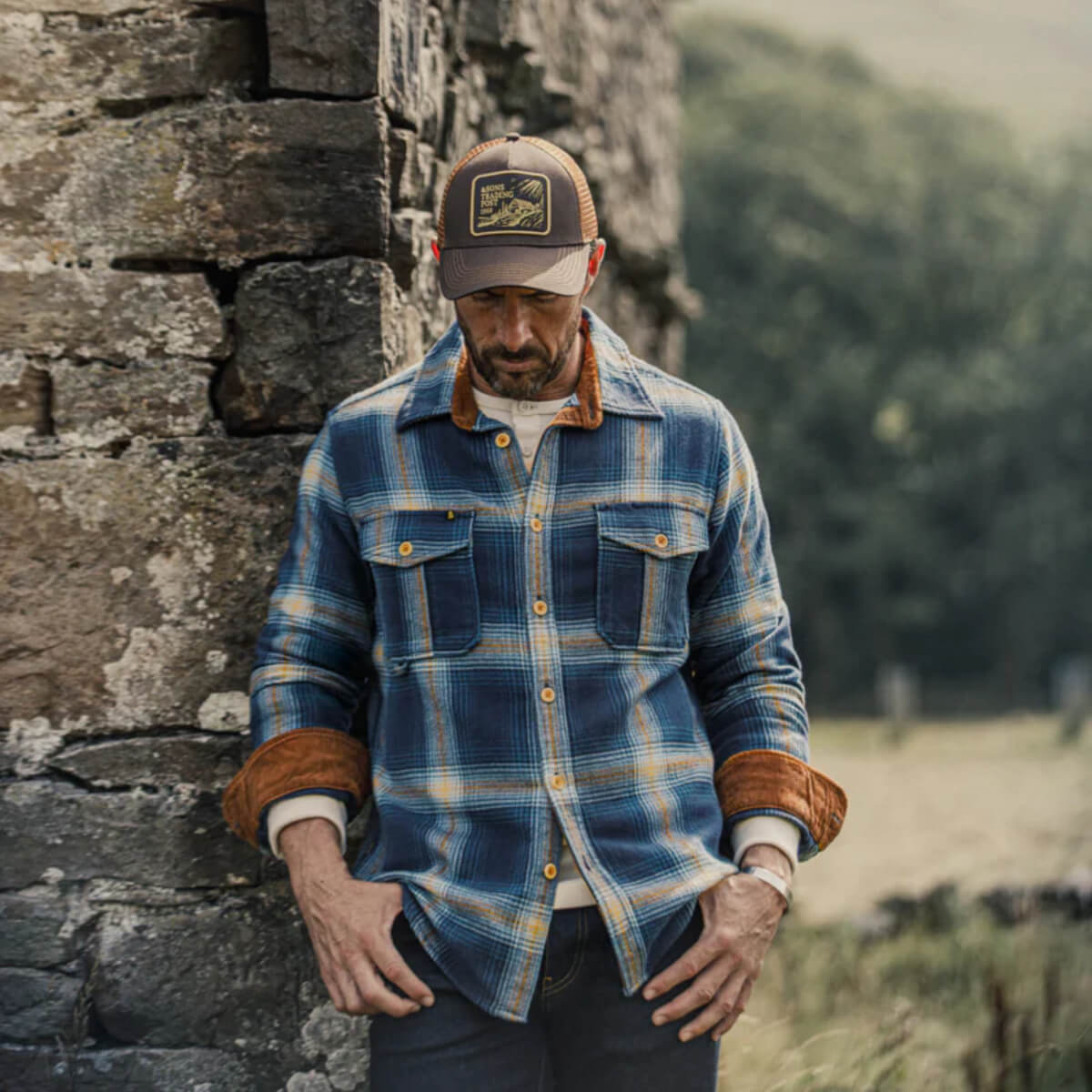 Man wearing a blue plaid shirt and cap leaning against a stone wall with a natural background
