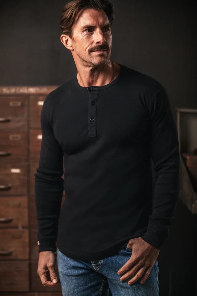 A man wearing a black long-sleeve henley shirt and blue jeans, standing in a dimly lit room with wooden cabinets in the background.