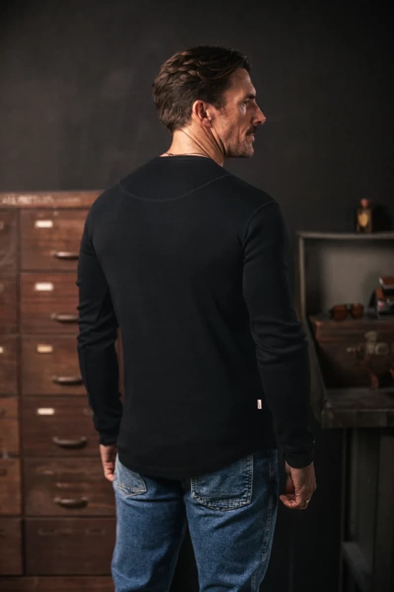 A man wearing a black long-sleeve crewneck shirt and blue jeans, shown from the back in a dimly lit room with wooden cabinets.