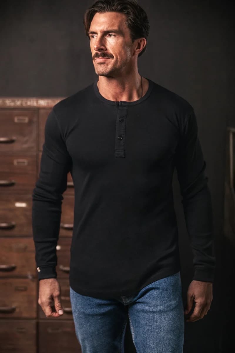 A man wearing a black long-sleeve henley shirt and blue jeans, standing in front of a dark background with wooden drawers.