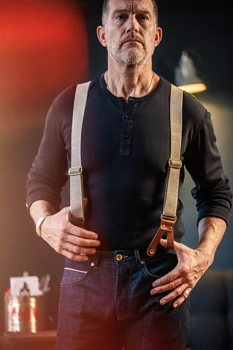 A man wearing beige canvas suspenders with brown leather accents over a black henley shirt and dark jeans, standing in a dimly lit environment with warm red and yellow lighting.