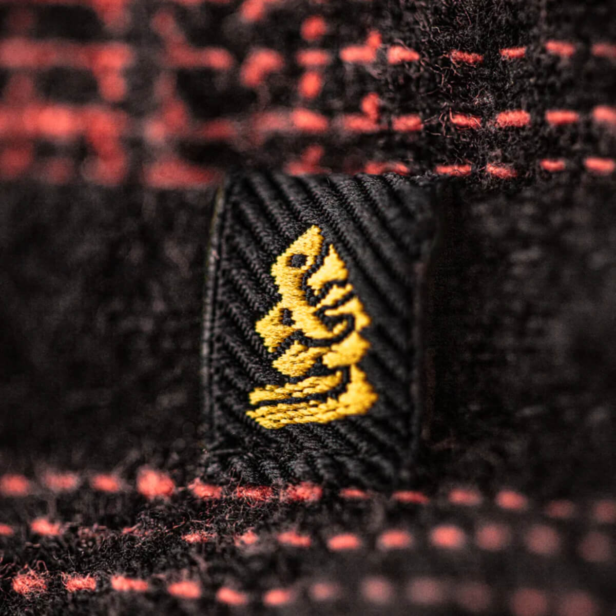 Close-up of a black fabric with a yellow embroidered logo on a red and black checkered background.
