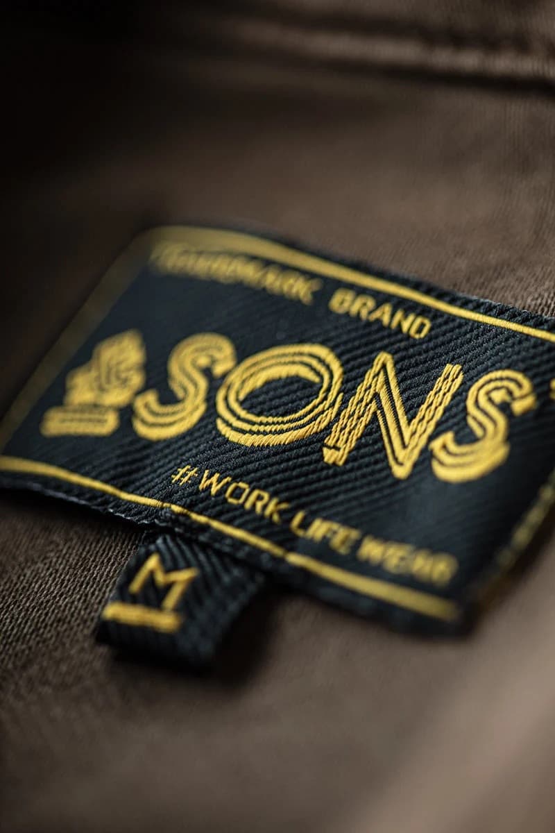 Close-up of a dark navy woven brand label with gold embroidery reading '&SONS' and '#WORKLIFEWEAR', attached to brown fabric.