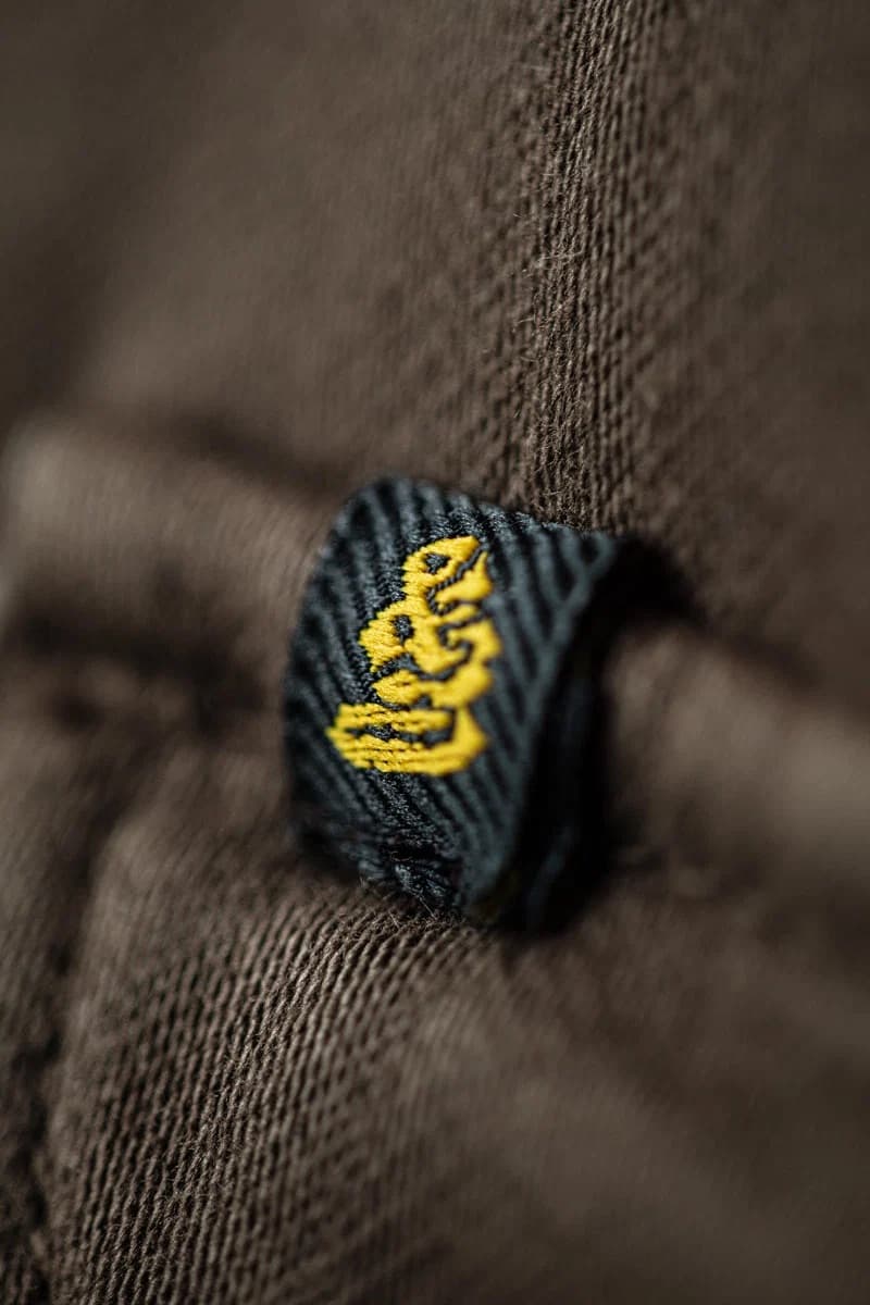 Close-up of a dark charcoal woven fabric tag with yellow embroidered stylized lettering, attached to a brown textured garment.