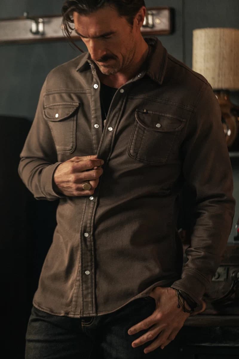 A man wearing a dark brown &SONS long-sleeve button-up Sunday shirt with two chest pockets, shown in a dimly lit indoor setting.
