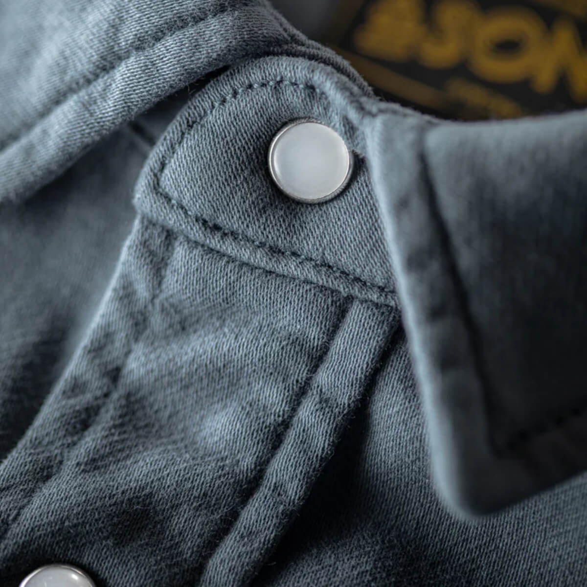 Close-up of a gray denim jacket with a button and part of a logo in the background.