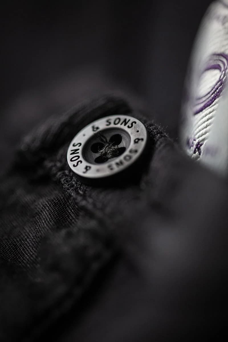 Close-up of a black textured fabric garment with a round silver-tone button labeled '&SONS' and a white patterned fabric partially visible on the right.