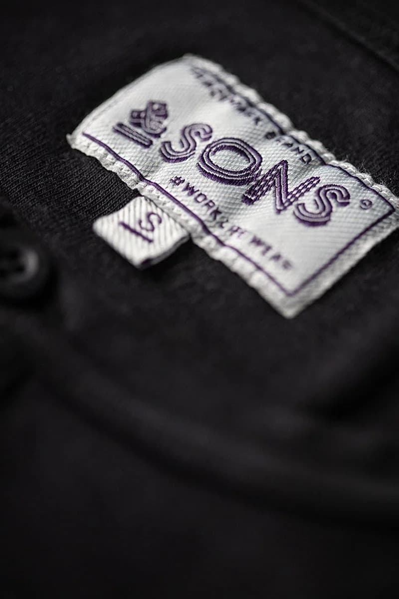 Close-up of a black knit garment with a sewn-on fabric label reading '&SONS' and 'WORK IT OUT' in purple embroidery.