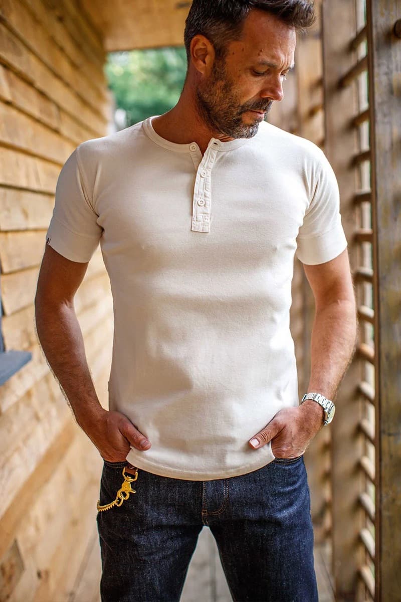 Man wearing an &SONS raw henley shirt and dark jeans standing against a wooden wall.