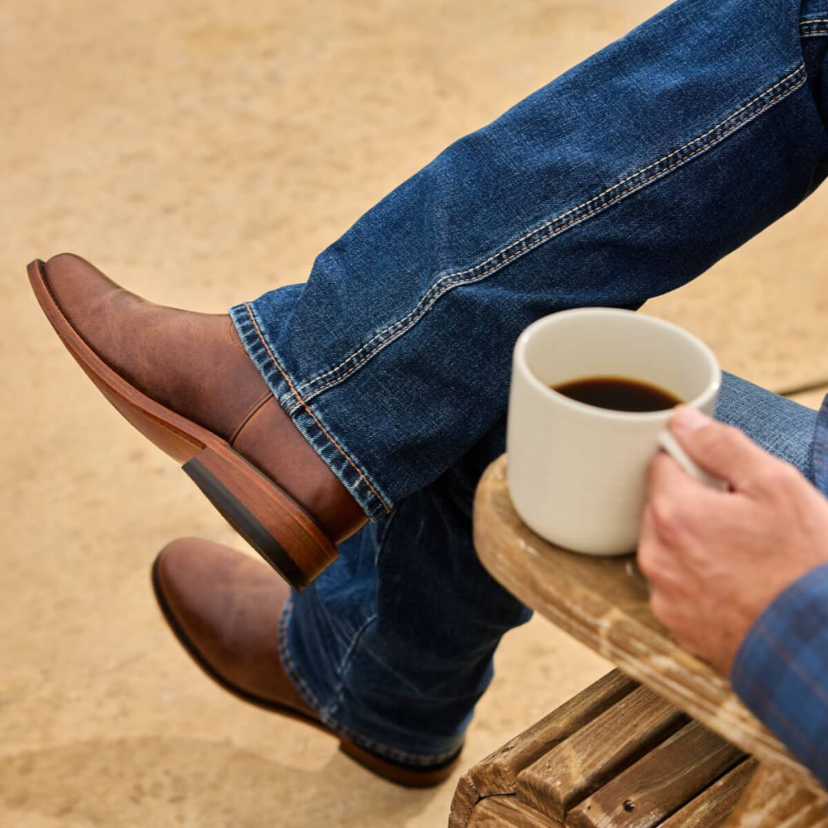 Person wearing brown leather shoes and blue jeans, holding a white mug with coffee on a wooden stool.