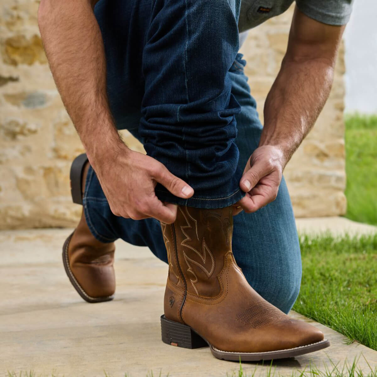 Person wearing brown cowboy boots with a stone wall and grass background