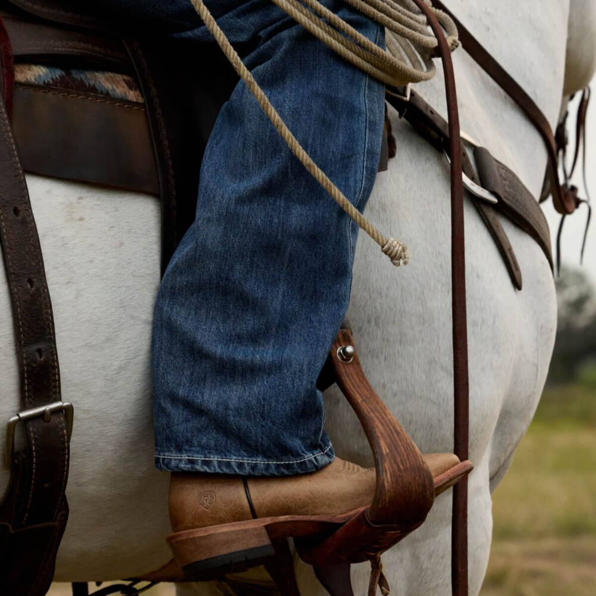 Close-up of a person wearing blue jeans and brown boots on a horse, with a blurred natural background.