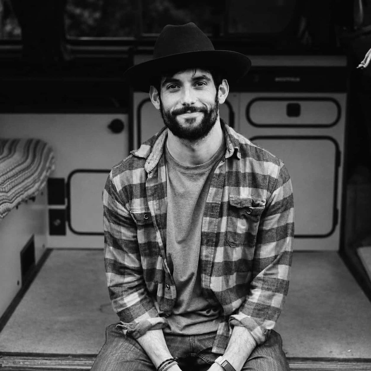 A man wearing a black Stetson Open Road vhat, a brown and gray plaid flannel shirt over a gray t-shirt, and jeans, sitting inside what appears to be a camper van or RV.