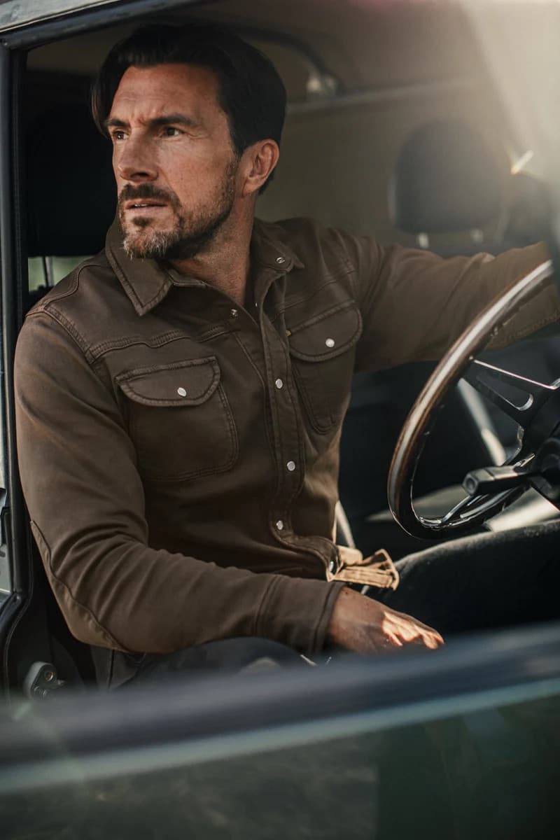 A man with a beard sitting in the driver's seat of a vehicle, wearing a dark brown long-sleeve western-style button-up shirt.