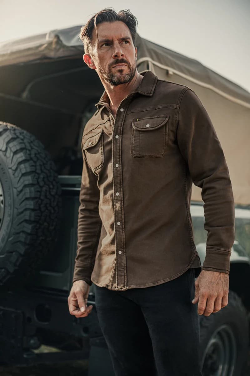 A man wearing a &SONS long-sleeve button-up Sunday shirt  and black pants, standing outdoors in front of a vehicle with a spare tire visible.