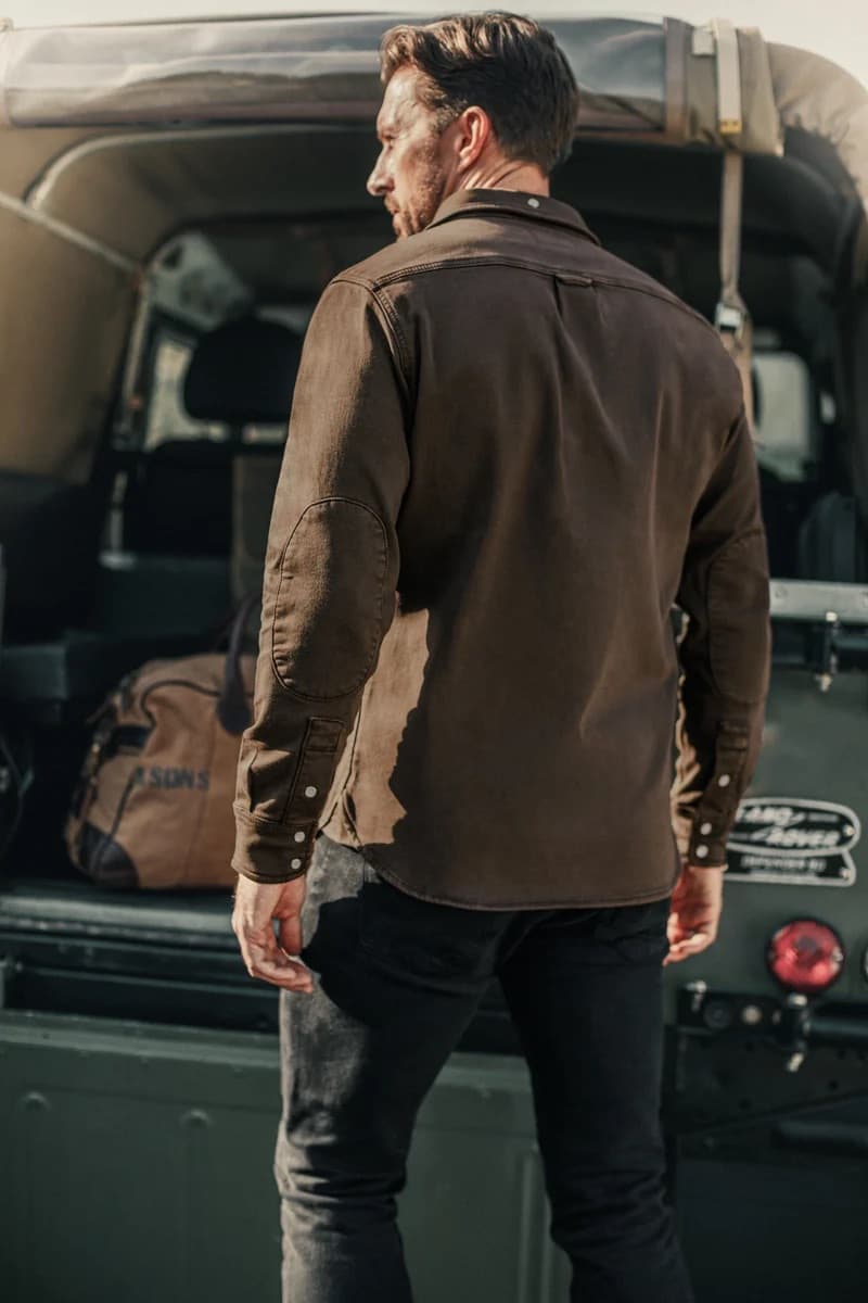 A man wearing a &SONS long-sleeve button-up Sunday shirt  and dark jeans, standing in front of the open trunk of a vintage green Land Rover with a brown duffel bag in the background.