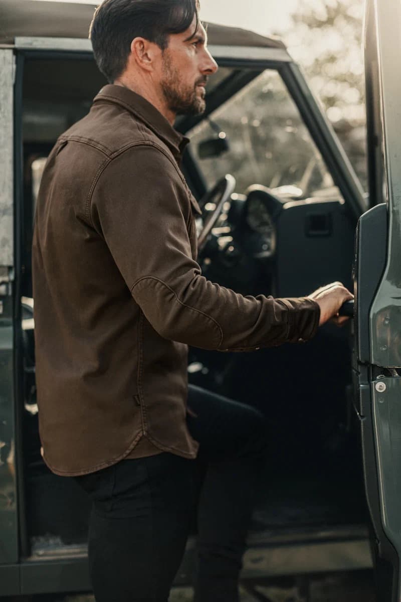 A man wearing a &SONS long-sleeve button-up Sunday shirt and black pants stands by the open door of a dark green vehicle, reaching in.