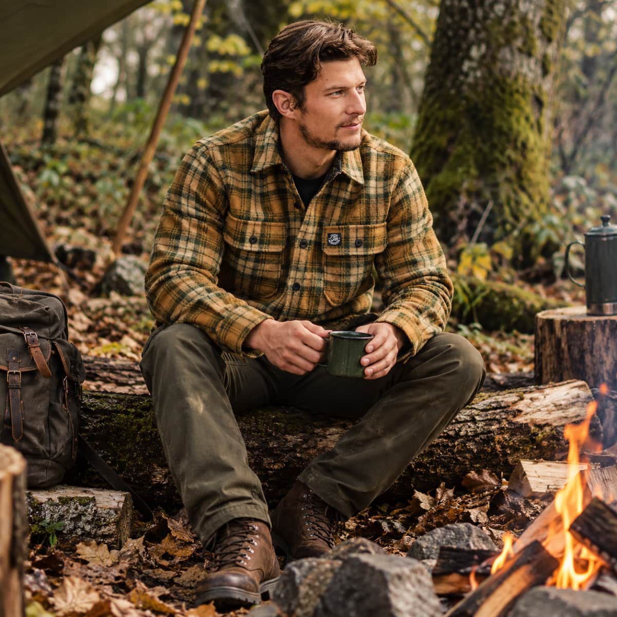 A man sits outdoors by a campfire wearing a brown and tan plaid flannel shirt, dark pants, and brown boots, holding a dark mug in a wooded setting.