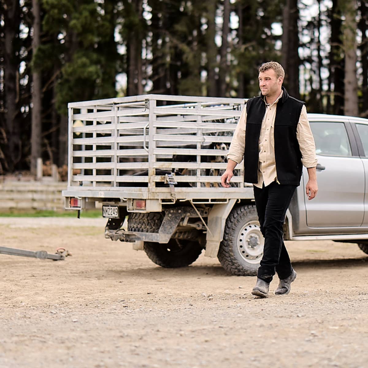 A man in a black vest and beige shirt walks beside a silver pickup truck with a metal cage bed on a dirt ground, with a forest in the background.