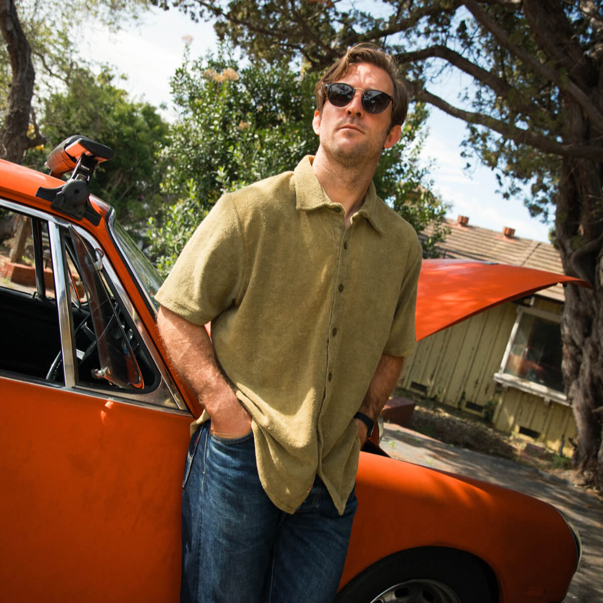 Man standing next to a vintage orange car with trees and a house in the background