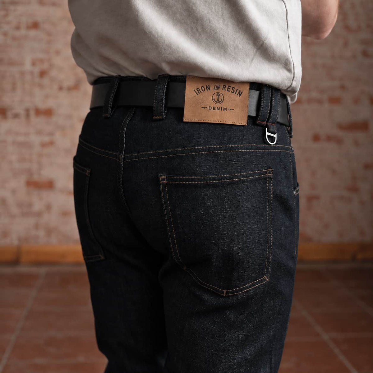 Rear view of a person wearing dark indigo denim jeans with a black belt and a tan leather patch that reads 'IRON RESIN DENIM'. The background shows a rustic brick wall and tiled floor.