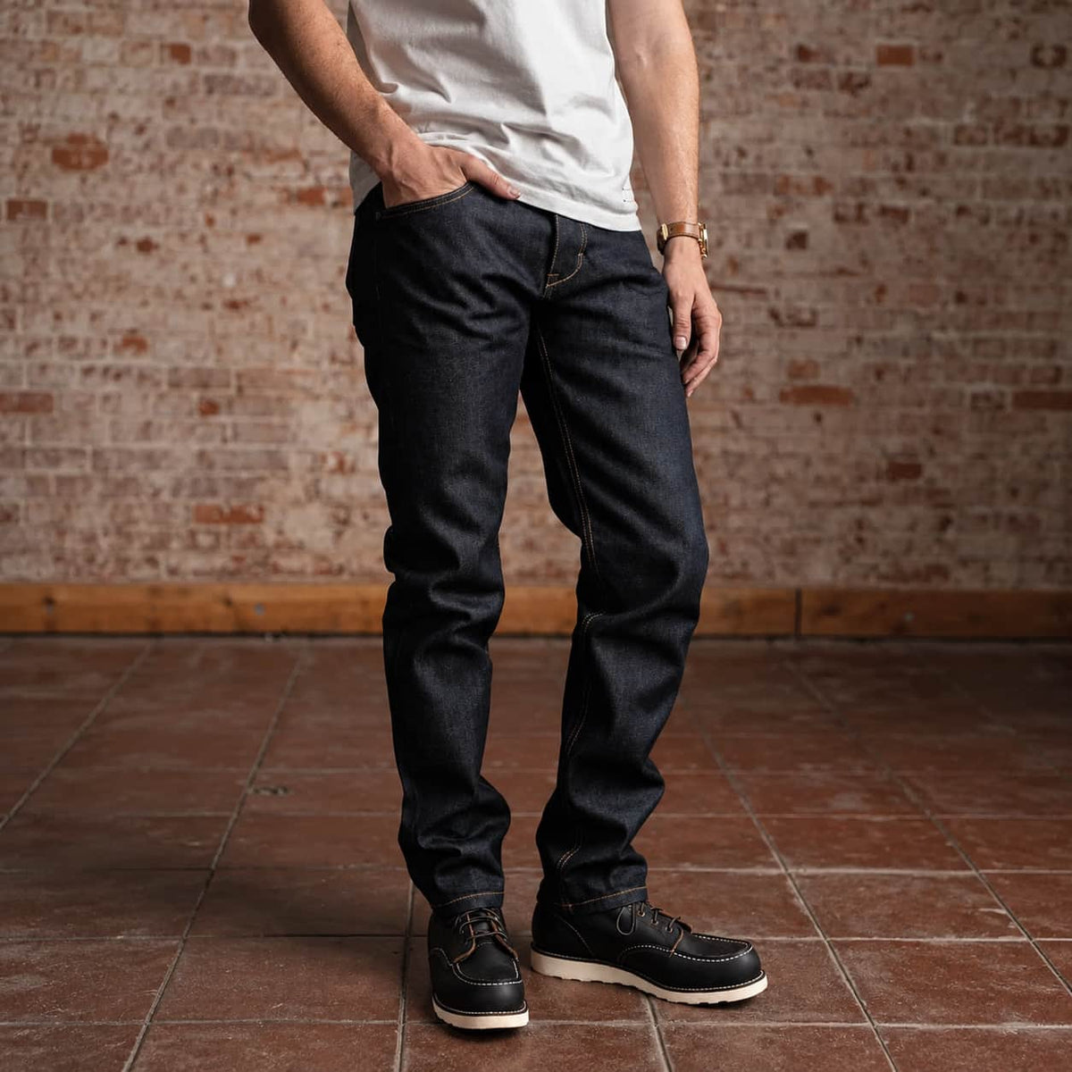 A man wearing dark indigo slim-fit jeans, a white t-shirt, and black leather shoes, standing in front of a rustic brick wall and tiled floor.