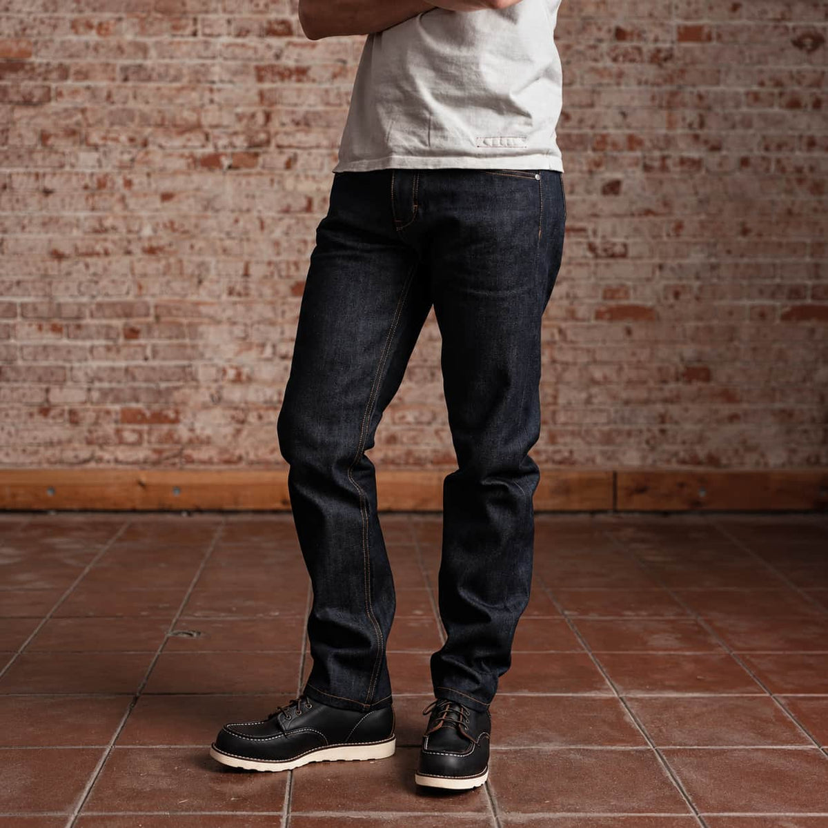A person wearing dark indigo straight-leg jeans, a light gray t-shirt, and black leather boat shoes, standing on a terracotta tiled floor against a rustic red brick wall.