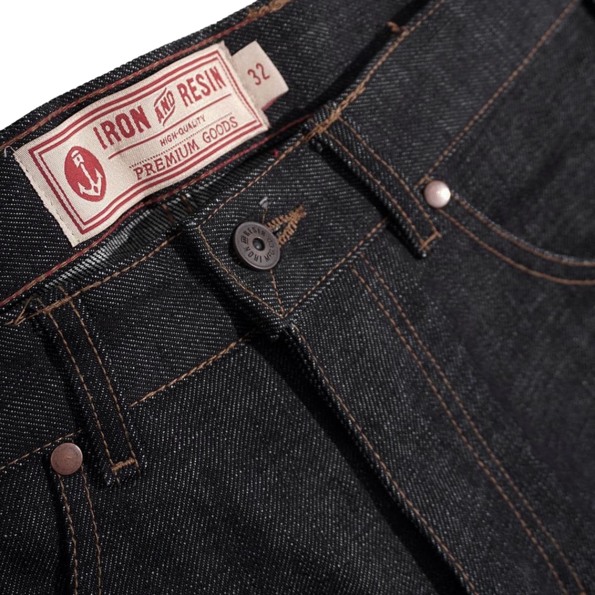Close-up of dark indigo denim jeans showing the waistband, button, rivets, and a sewn-in label that reads Iron Resin Premium Goods.