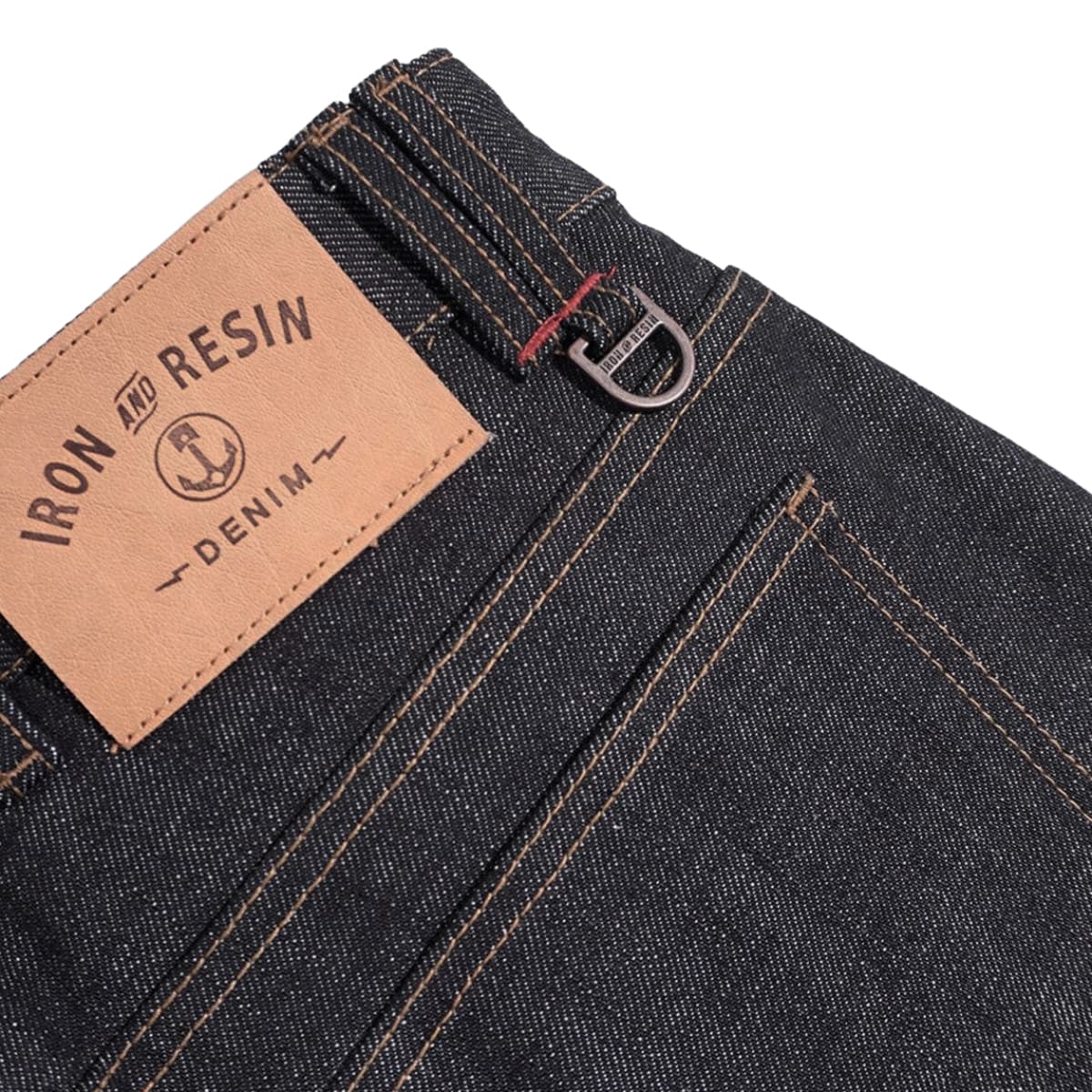 Close-up of dark indigo denim jeans showing the back waistband, belt loop, and a tan leather patch with the brand name 'IRON AND RESIN DENIM'.