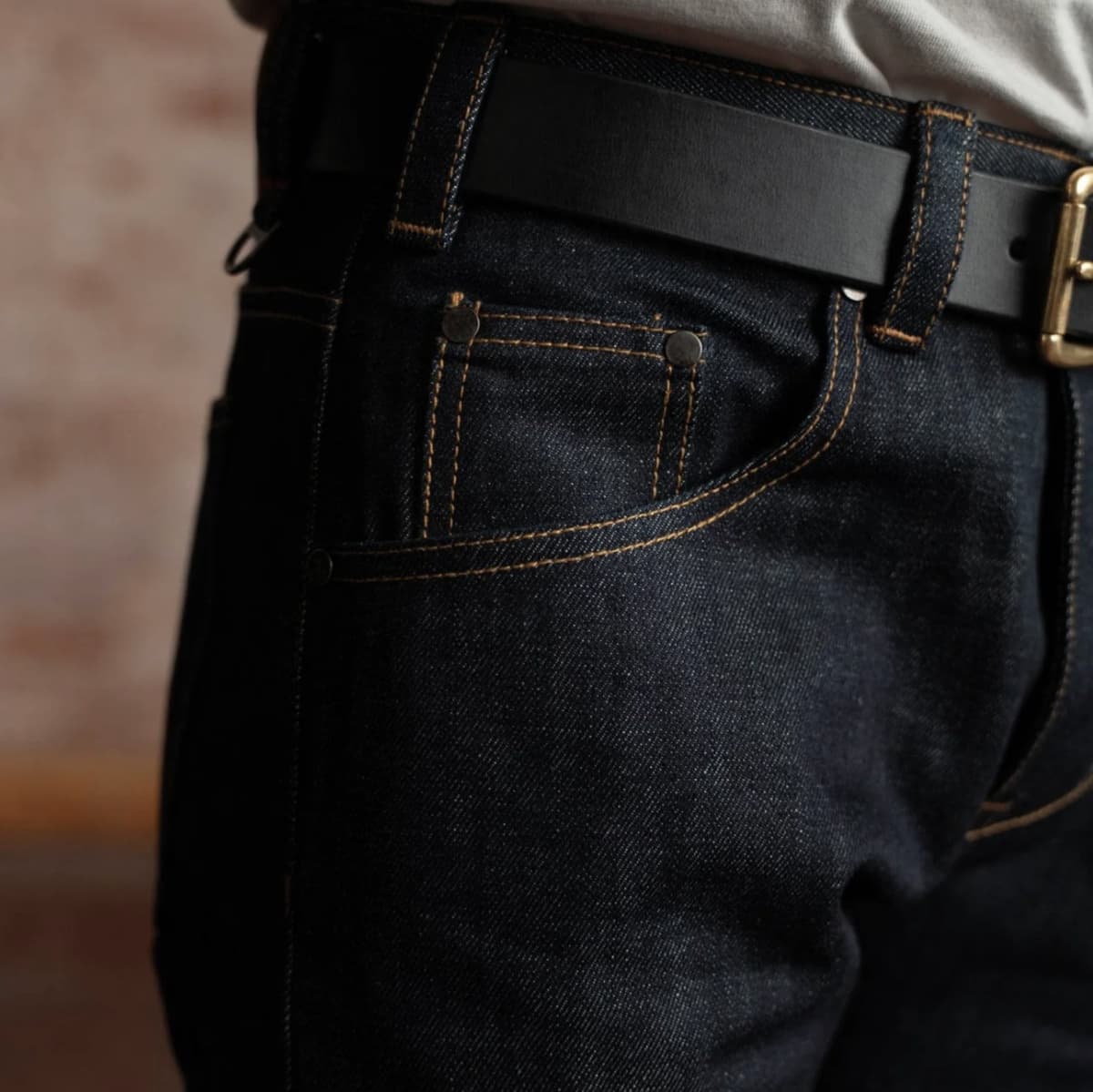 Close-up of a person wearing dark indigo denim jeans with contrast tan stitching, a black leather belt, and a gold-tone buckle, cropped from the waist down against a softly blurred indoor background.