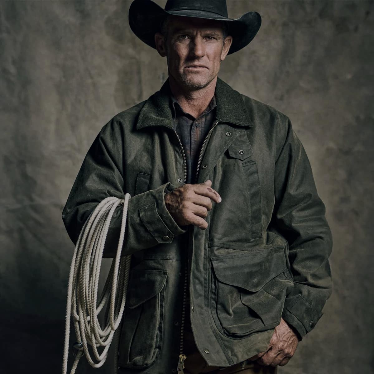 A man wearing a black cowboy hat and a Filson Tin Cloth Field Jacket, holding a coiled white rope against a textured dark grey studio backdrop.
