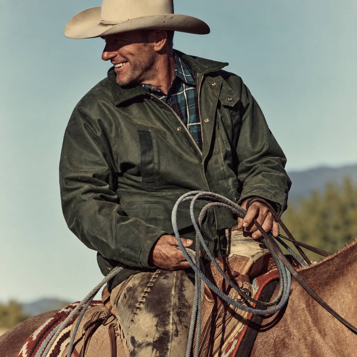 A smiling cowboy wearing a Filson Tin Cloth Field Jacket plaid shirt, and beige cowboy hat, sitting on a horse outdoors and holding a coiled lasso.