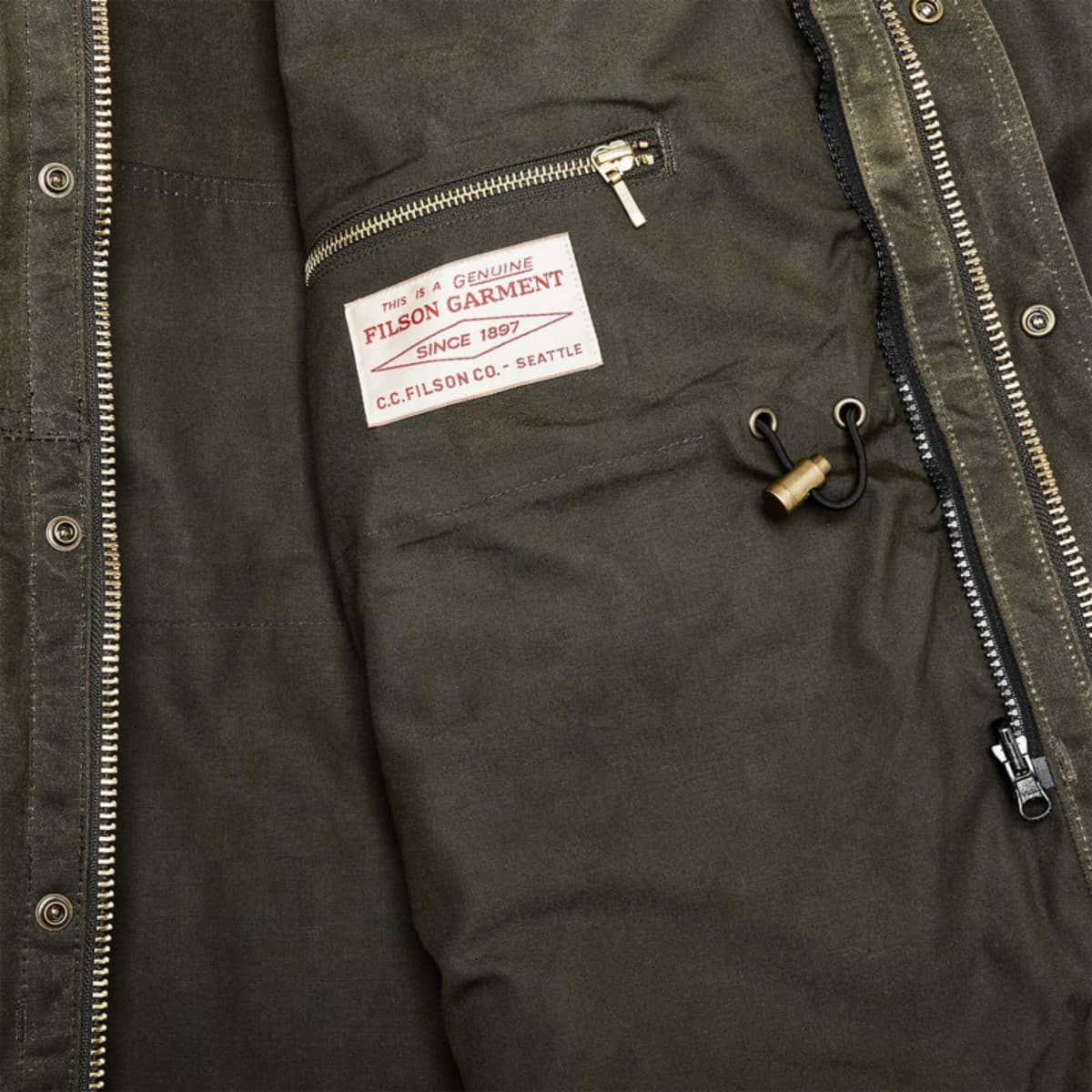 Close-up of a Filson Tin Cloth Field Jacket showing the inner pocket with a white Filson brand label, metal zipper, and drawstring detail.