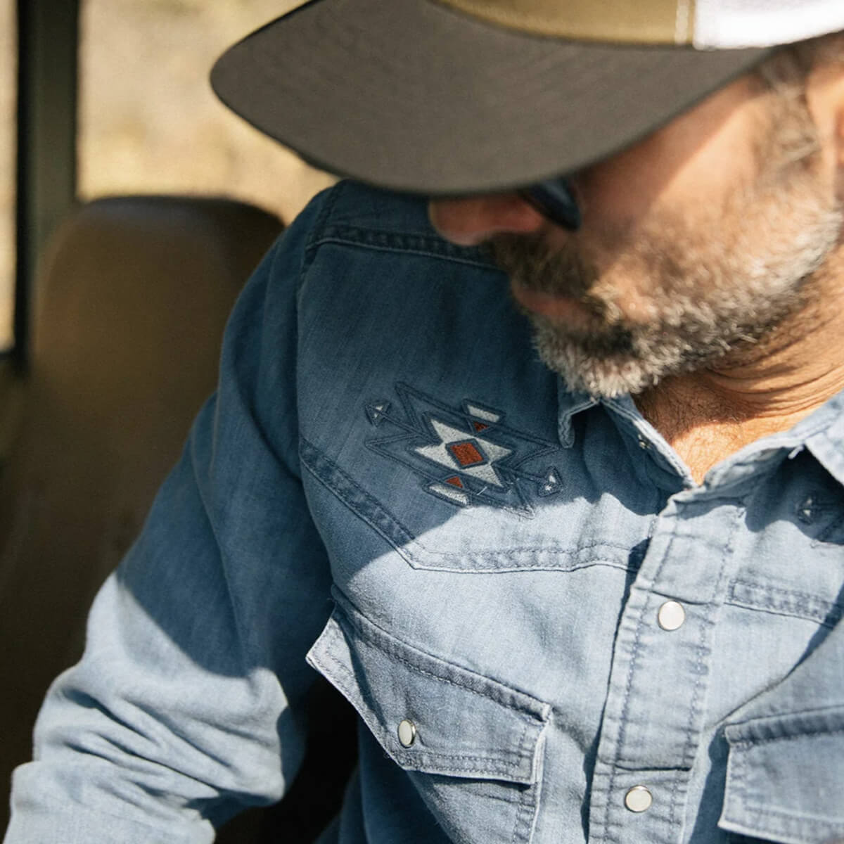 Man wearing a blue denim shirt with a logo and a cap, sitting inside a vehicle.