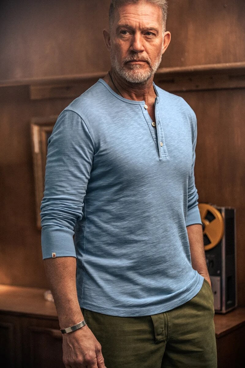 Man wearing a blue long-sleeve shirt and green pants standing against a wooden wall.