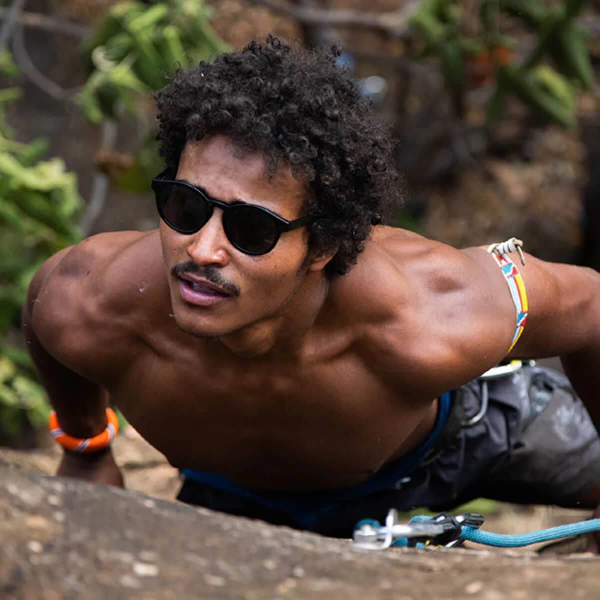 Man climbing a rock face wearing sunglasses and minimal clothing.
