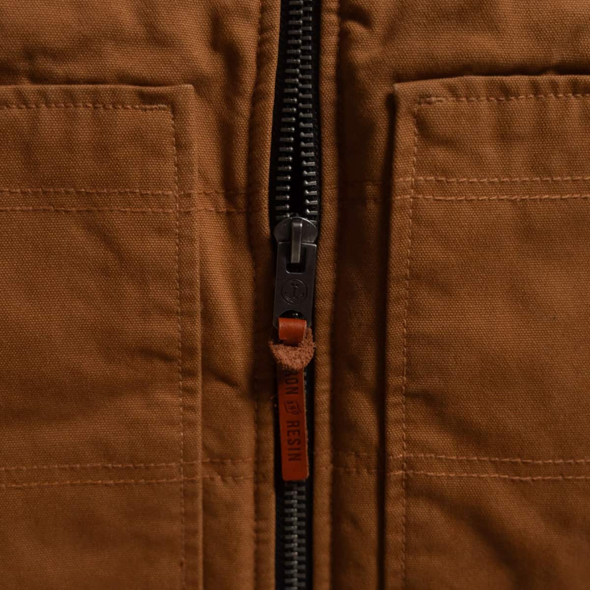 Close-up of a brown canvas jacket with a black zipper, featuring a brown leather pull tab with the brand name 'JAPAN BY DESIGN' printed vertically.