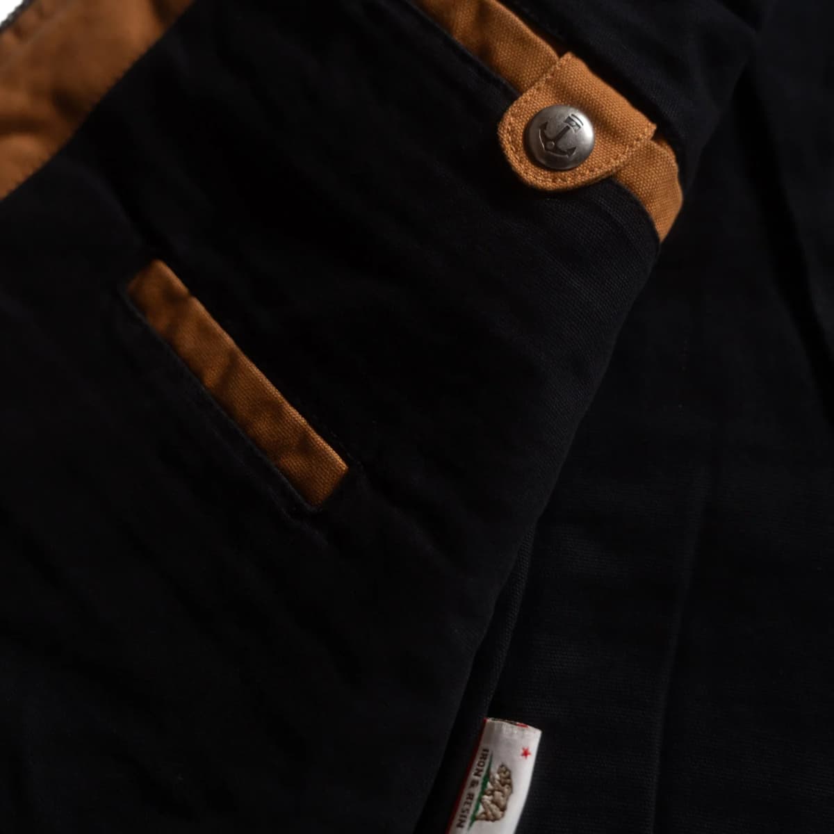 Close-up of a black canvas jacket with contrasting brown suede-like trim on the pocket and waistband, featuring a silver metal snap button.
