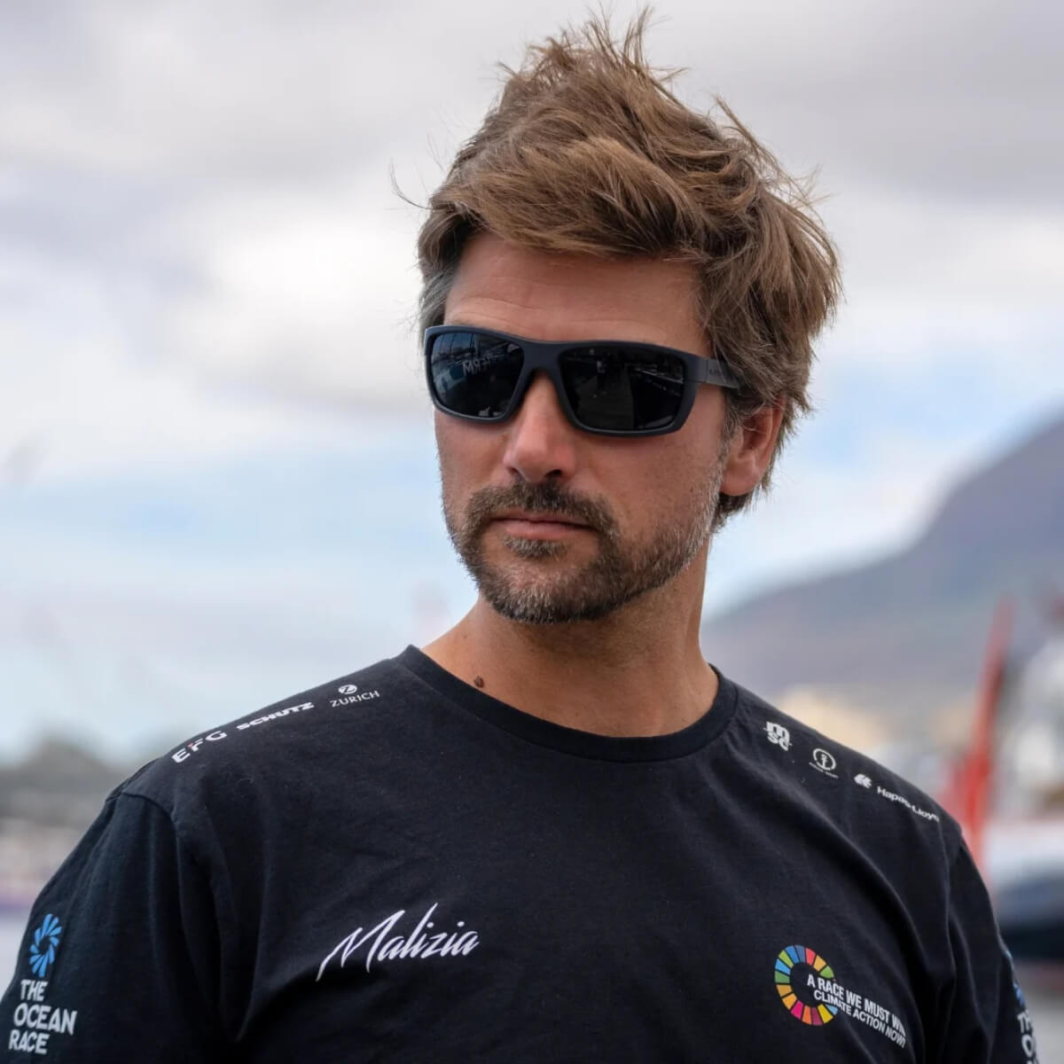Man wearing sunglasses and a black shirt with logos, outdoors.