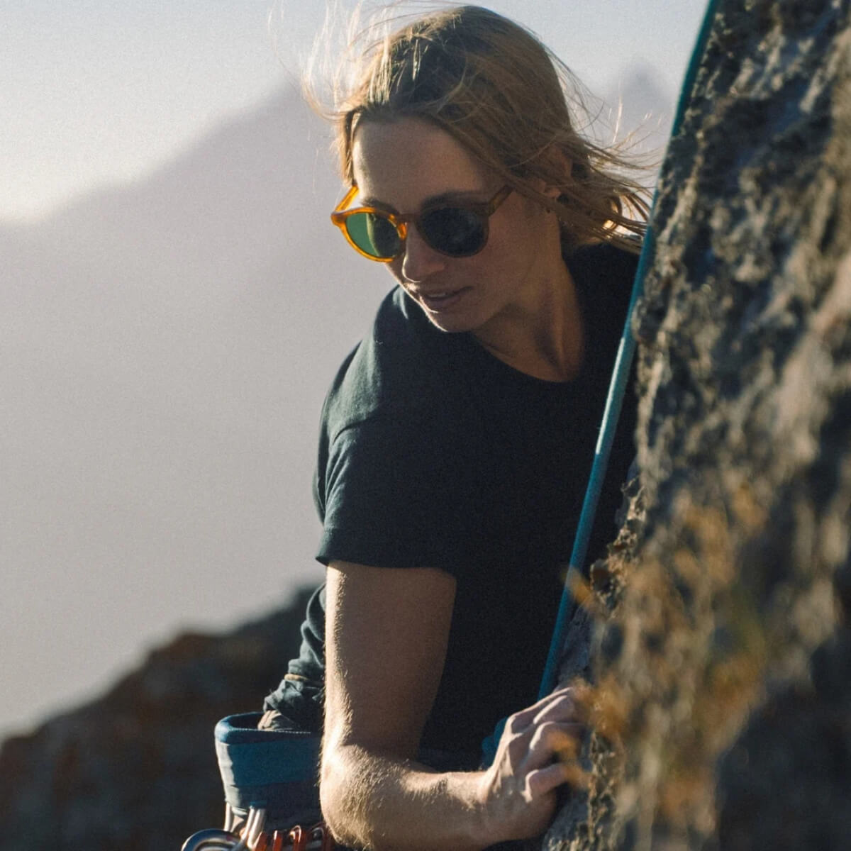 Person wearing sunglasses and a black shirt, holding a blue cup, with a mountainous background.